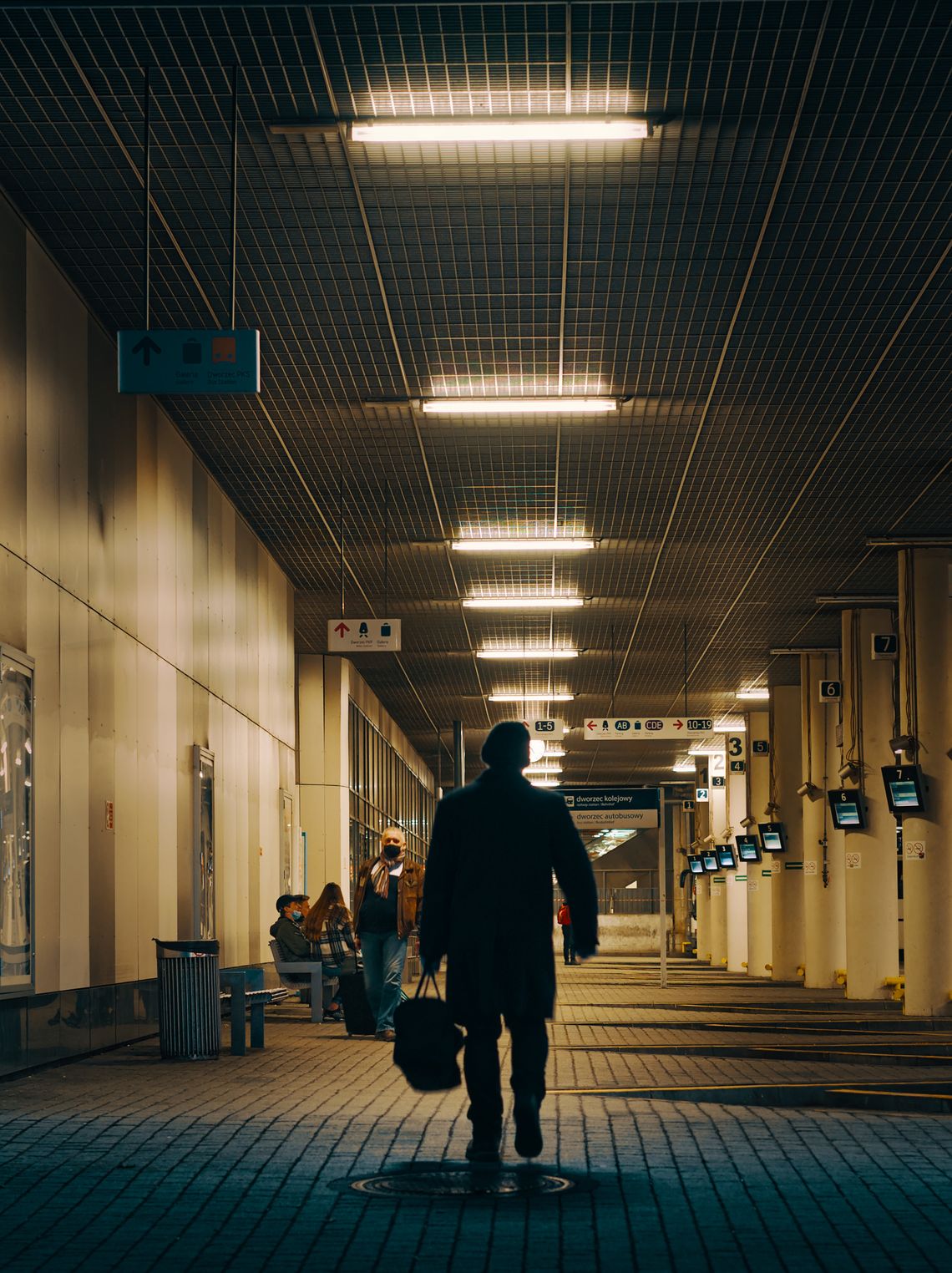 Dworzec PKS wieczorem, na pierwszym planie, na środku człowiek niosący bagaż w lewej ręce.