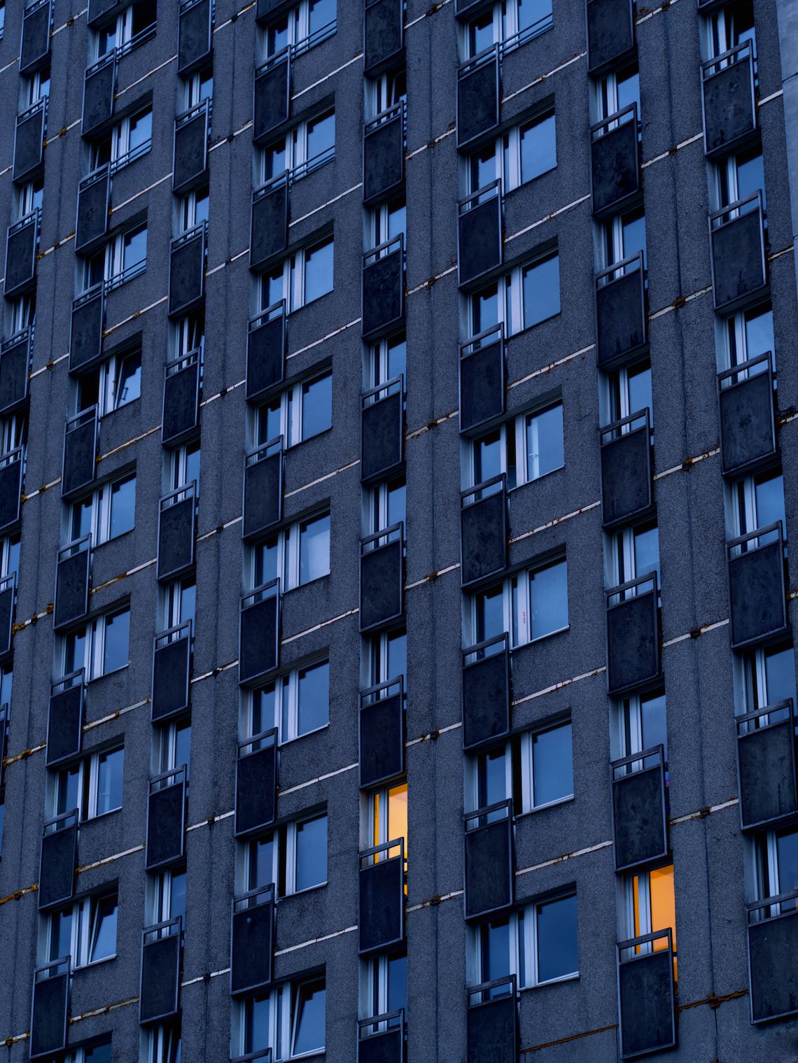 Zmierzch, ściana budynku z wielkiej płyty. Zapalone światło w dwóch oknach w prawym dolnym rogu.