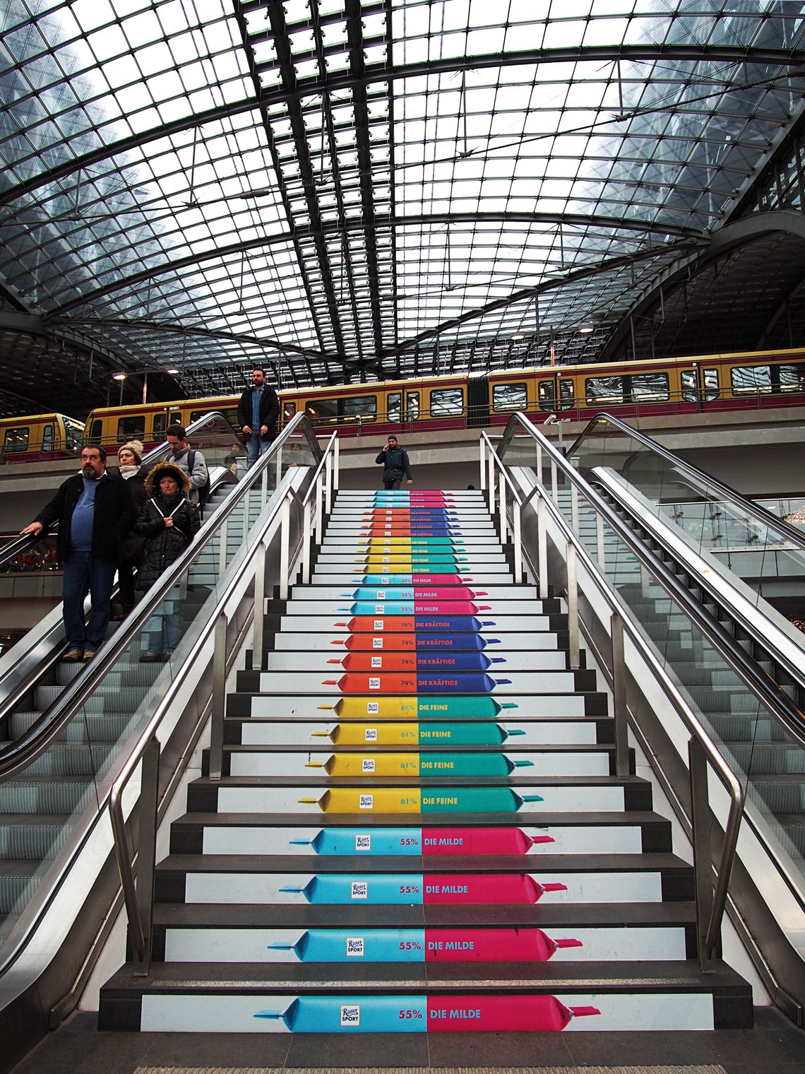 Schody na dworcu w Berlinie. Na schodkach kolorowe reklamy. U szczytu schodów mężczyzna. W tle kolejka U-Bahn.